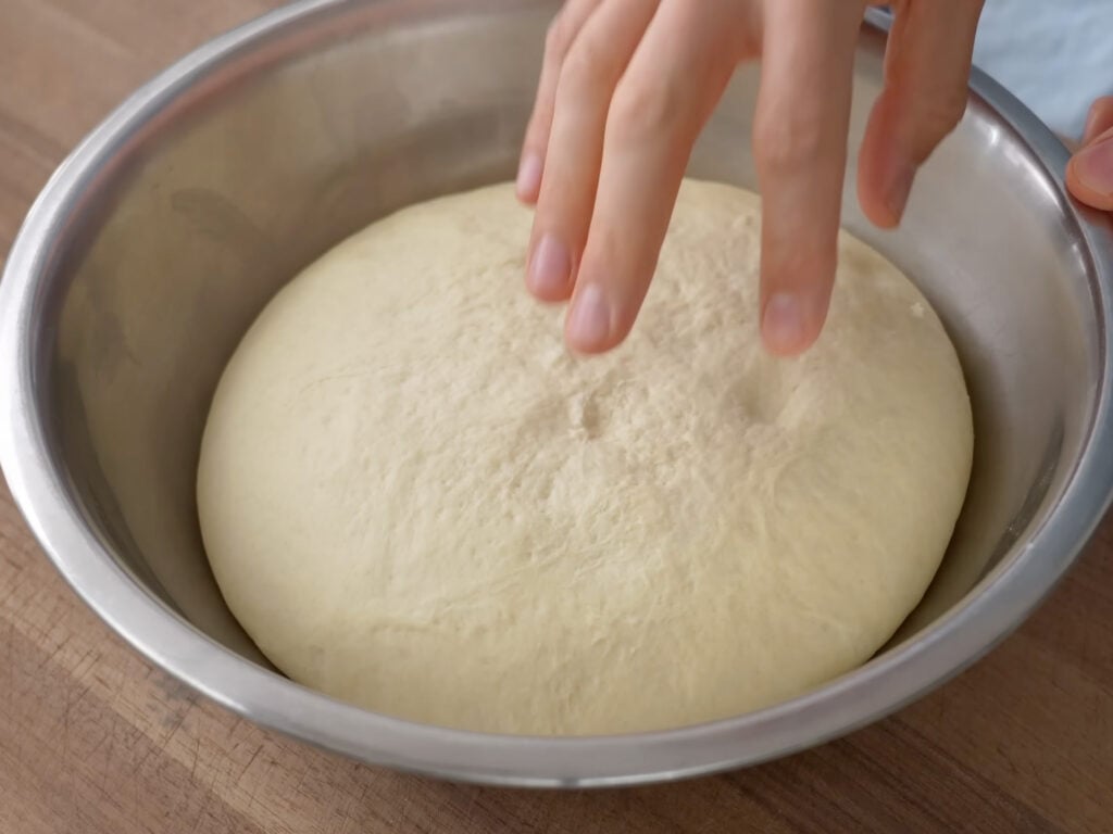 fingers poking proofed pizza dough in metal bowl