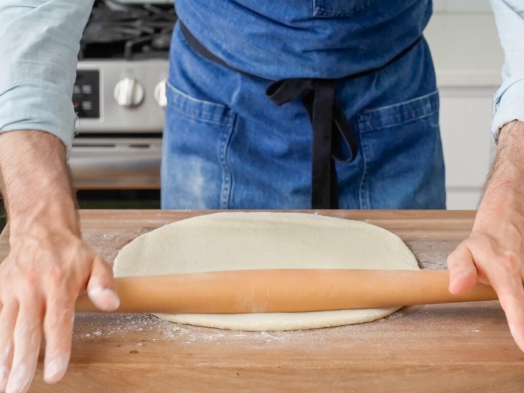 man in blue apron rolling out pizza dough