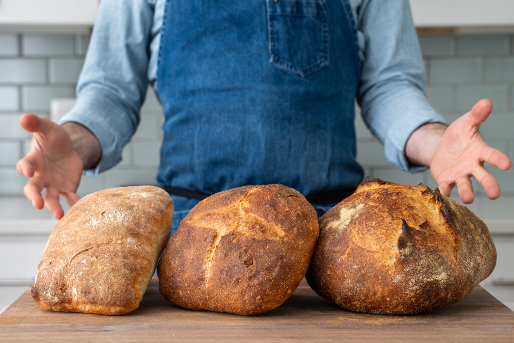 man in blue apron holding hands on either side of 3 loaves of bread on a cutting board
