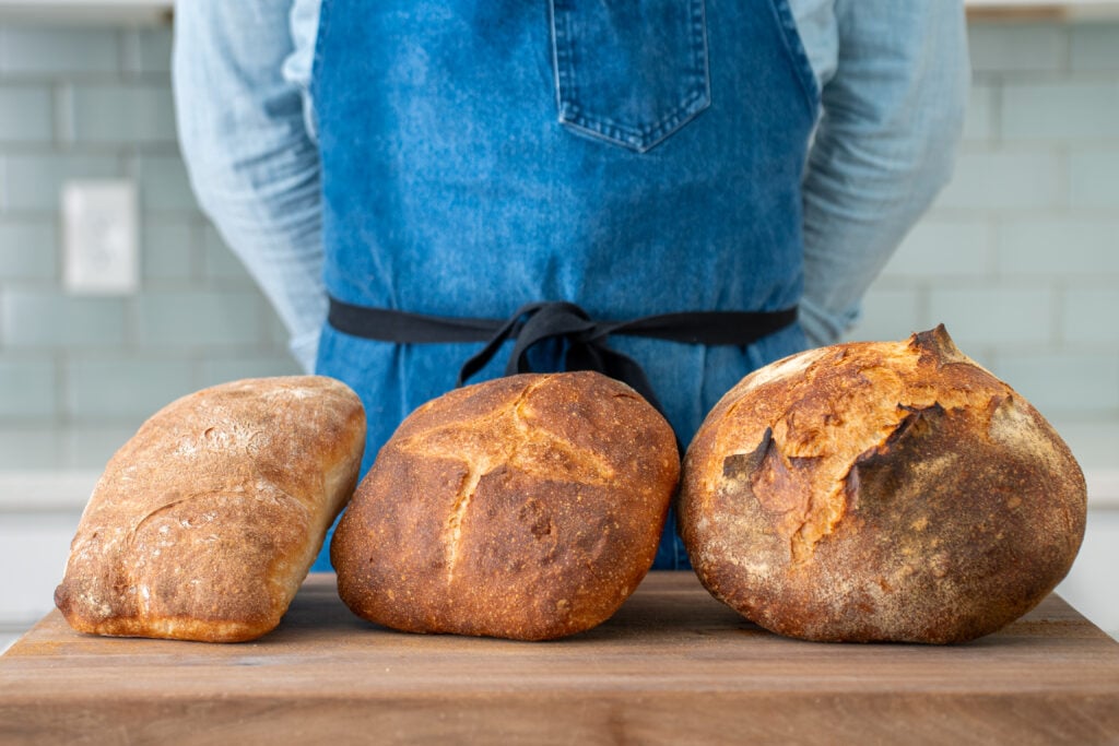 man in blue apron standing behind 3 loaves of bread