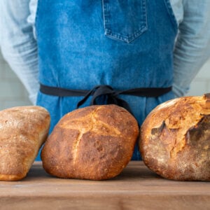 man in blue apron standing behind 3 loaves of bread