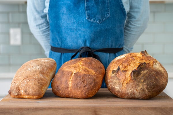 man in blue apron standing behind 3 loaves of bread