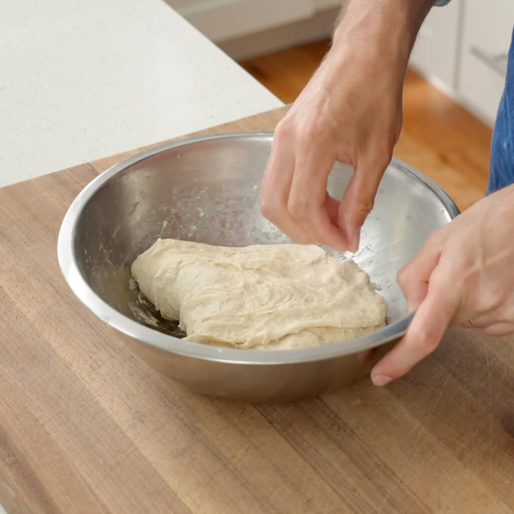 hand performing strength building fold on dough in metal bowl