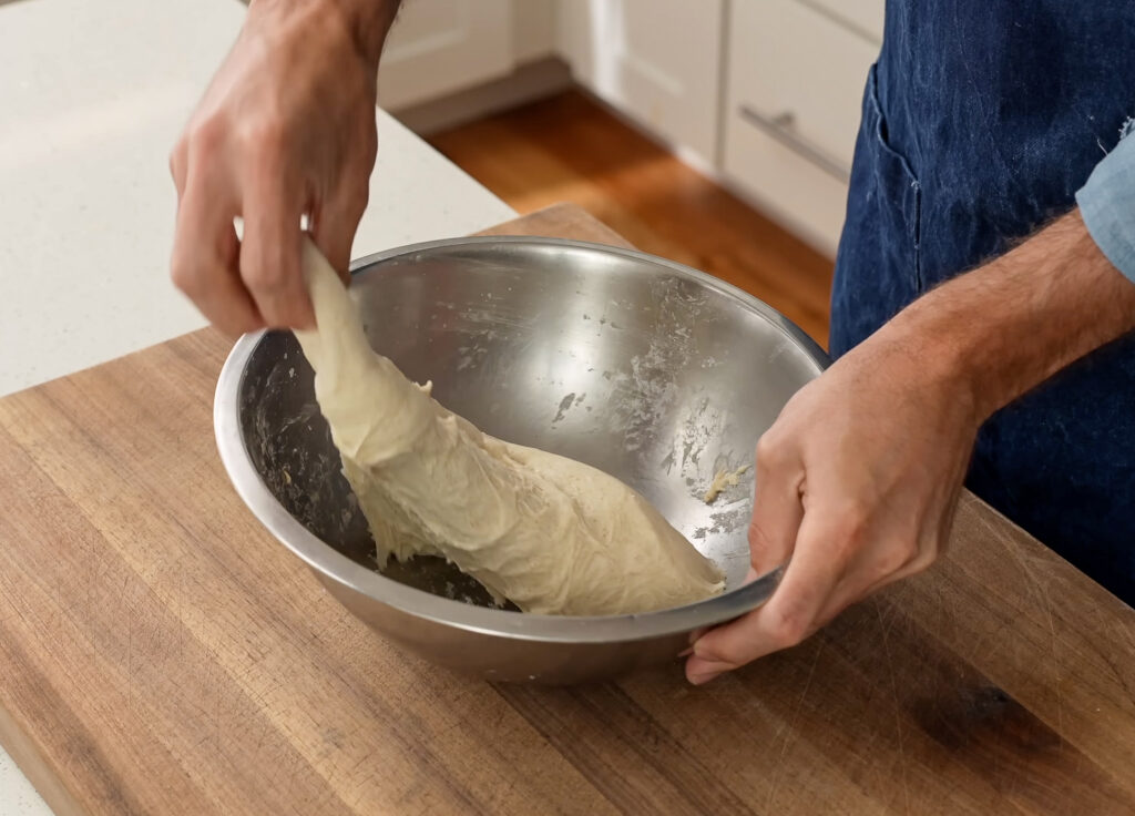 man pulling out and folding over dough in metal bowl