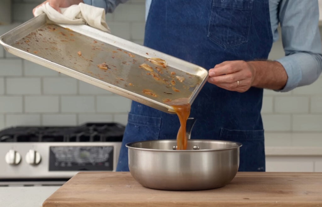 man pouring brown drippings from sheet pan into sauce pan