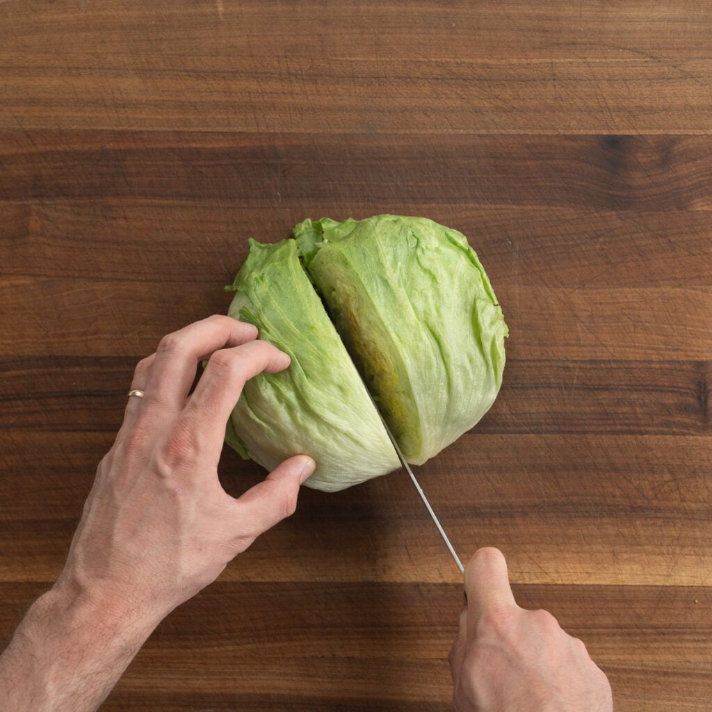 cutting head of iceberg lettuce in half on a wooden cutting board