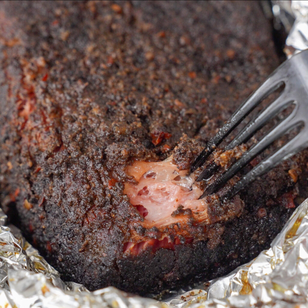 close up of cooked pastrami in foil being pulled apart with black fork