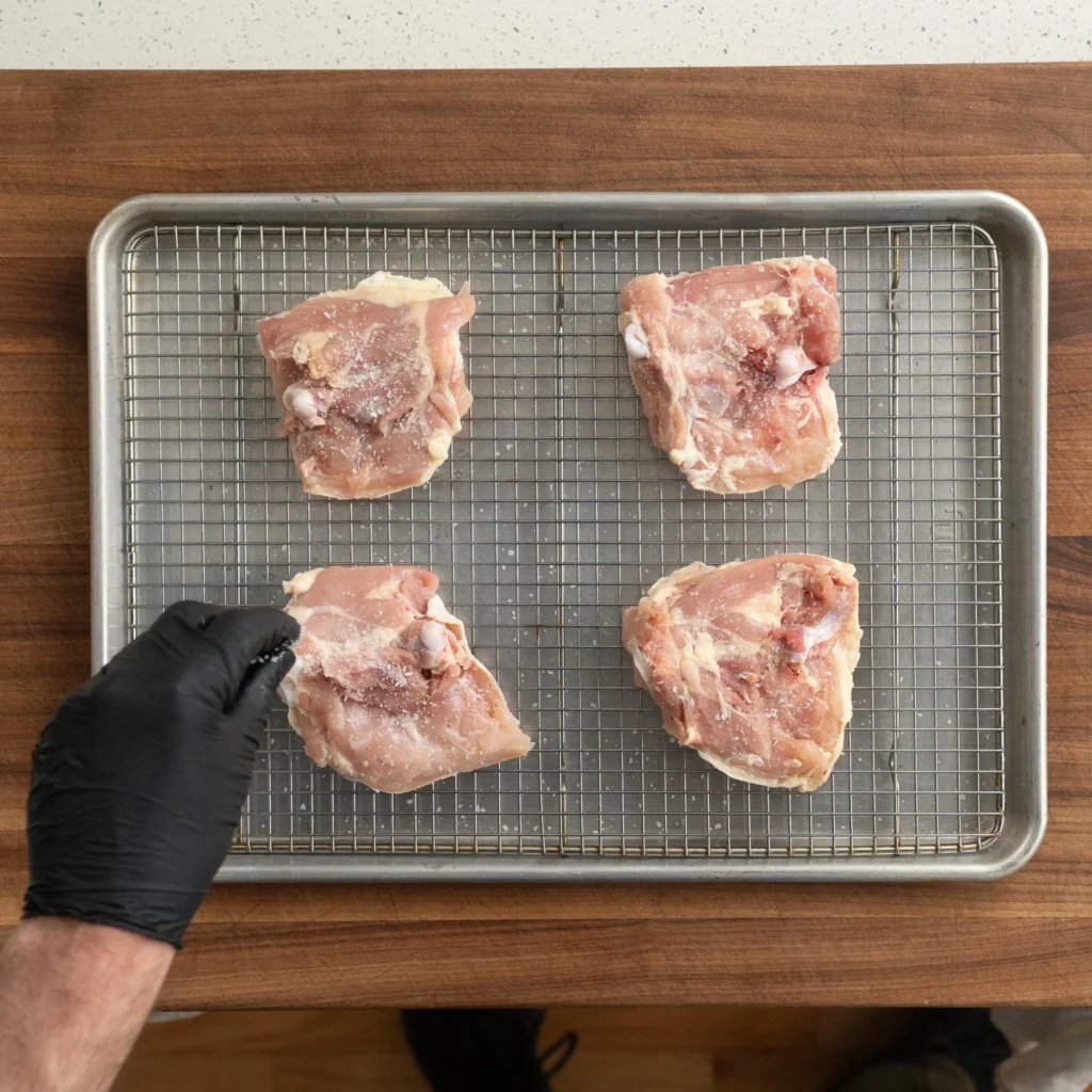 black gloved hand salting chicken thighs on wire rack
