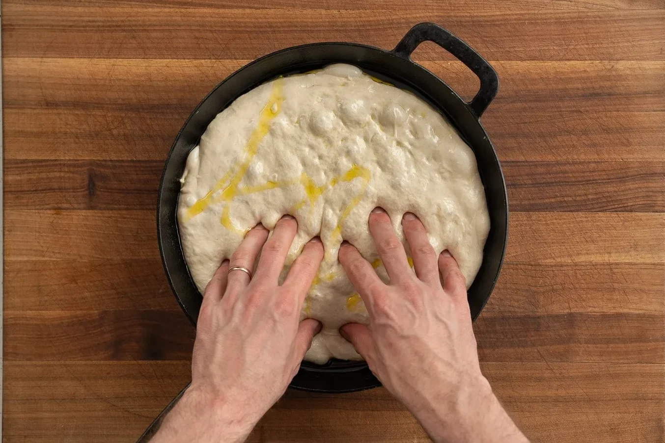 dimpling focaccia dough with fingertips in a cast iron pan