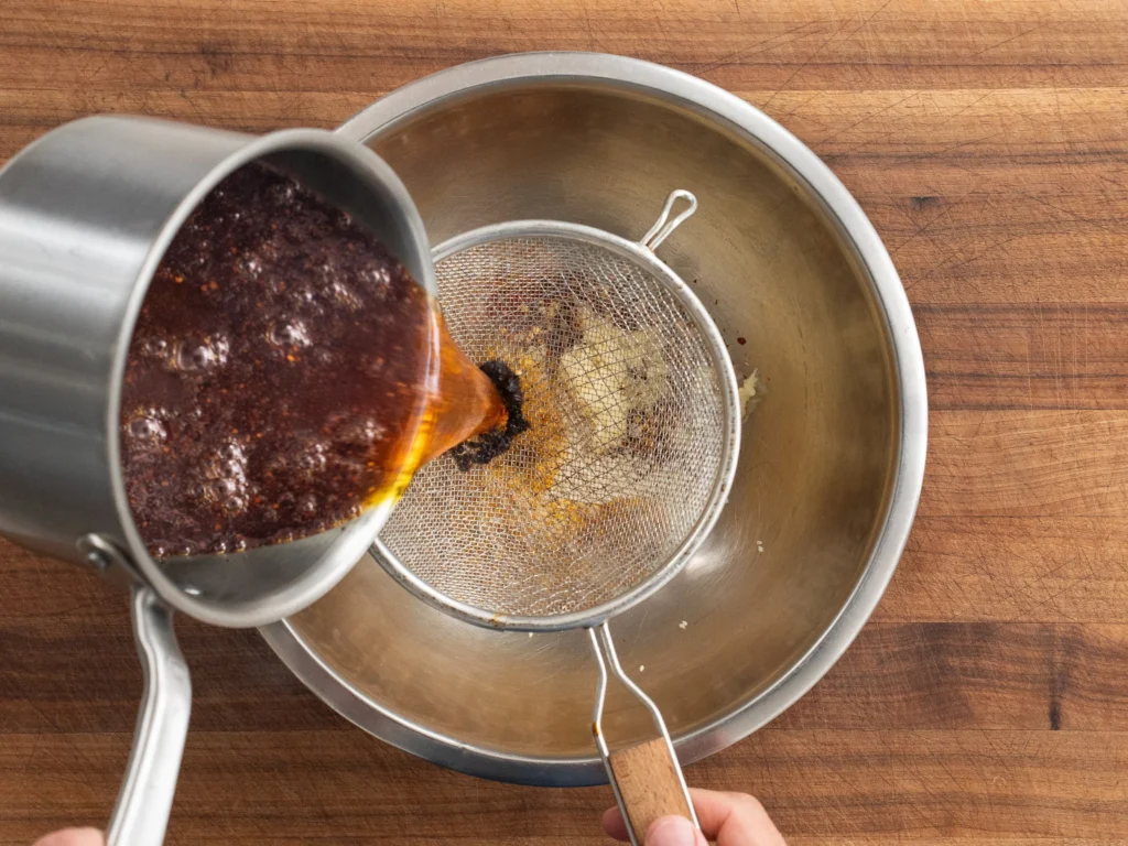 pouring wonton chili oil through a strainer into a metal bowl