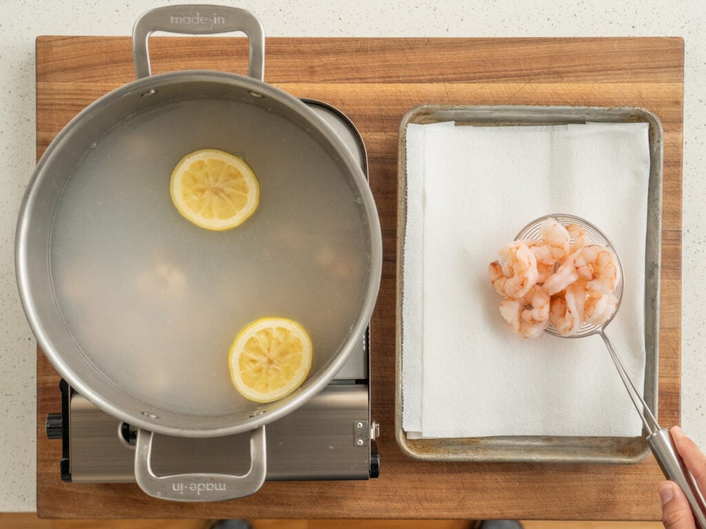 pot of cooked shrimp in water on the right with spider strainer setting cooked shrimp on paper towel lined tray on right