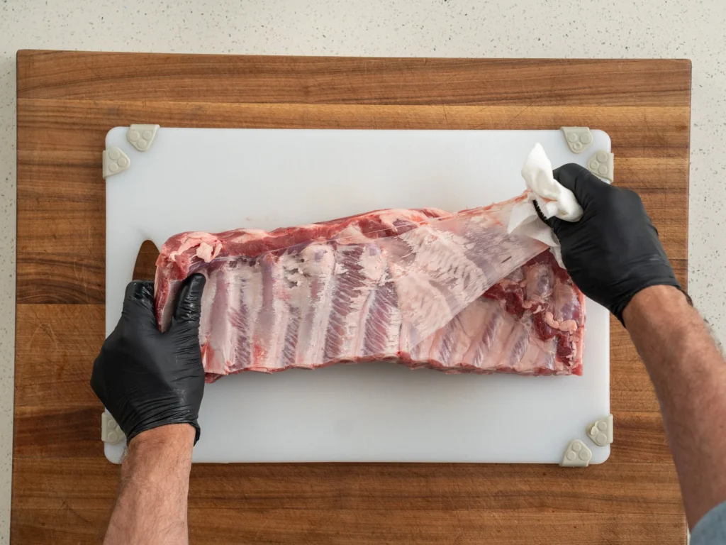 black gloved hands pulling pork membrane off rack of ribs