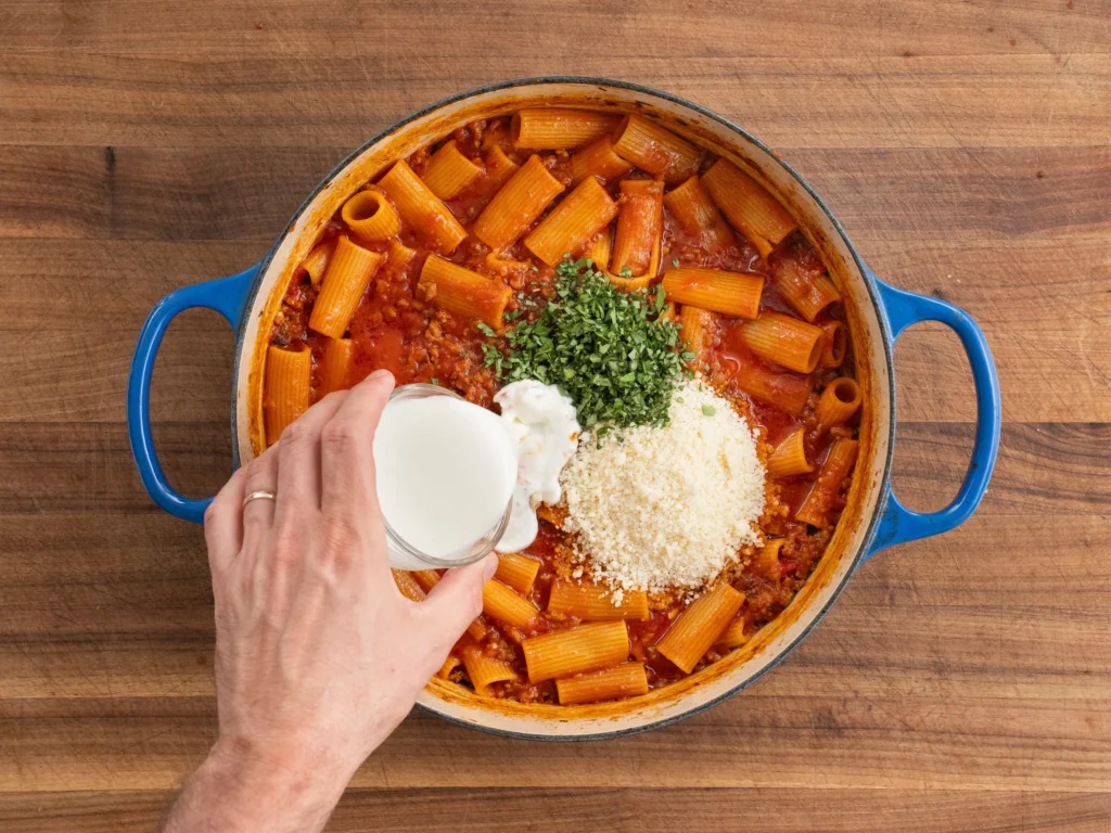 hand pouring heavy cream into a dutch oven filled with rigatoni pasta basil and parmesan