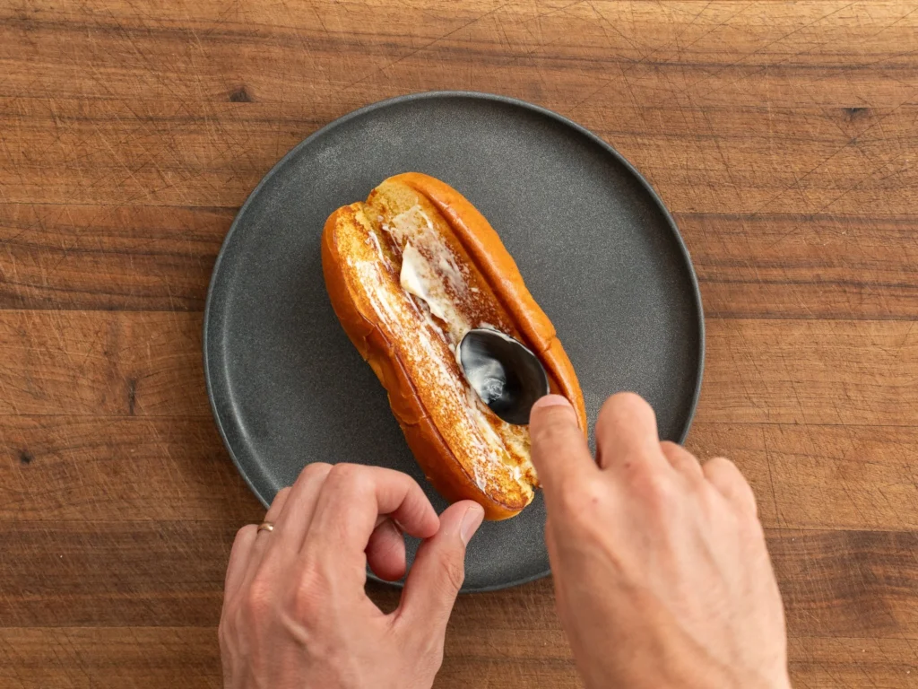 spreading mayonnaise in hot dog bun with black spoon