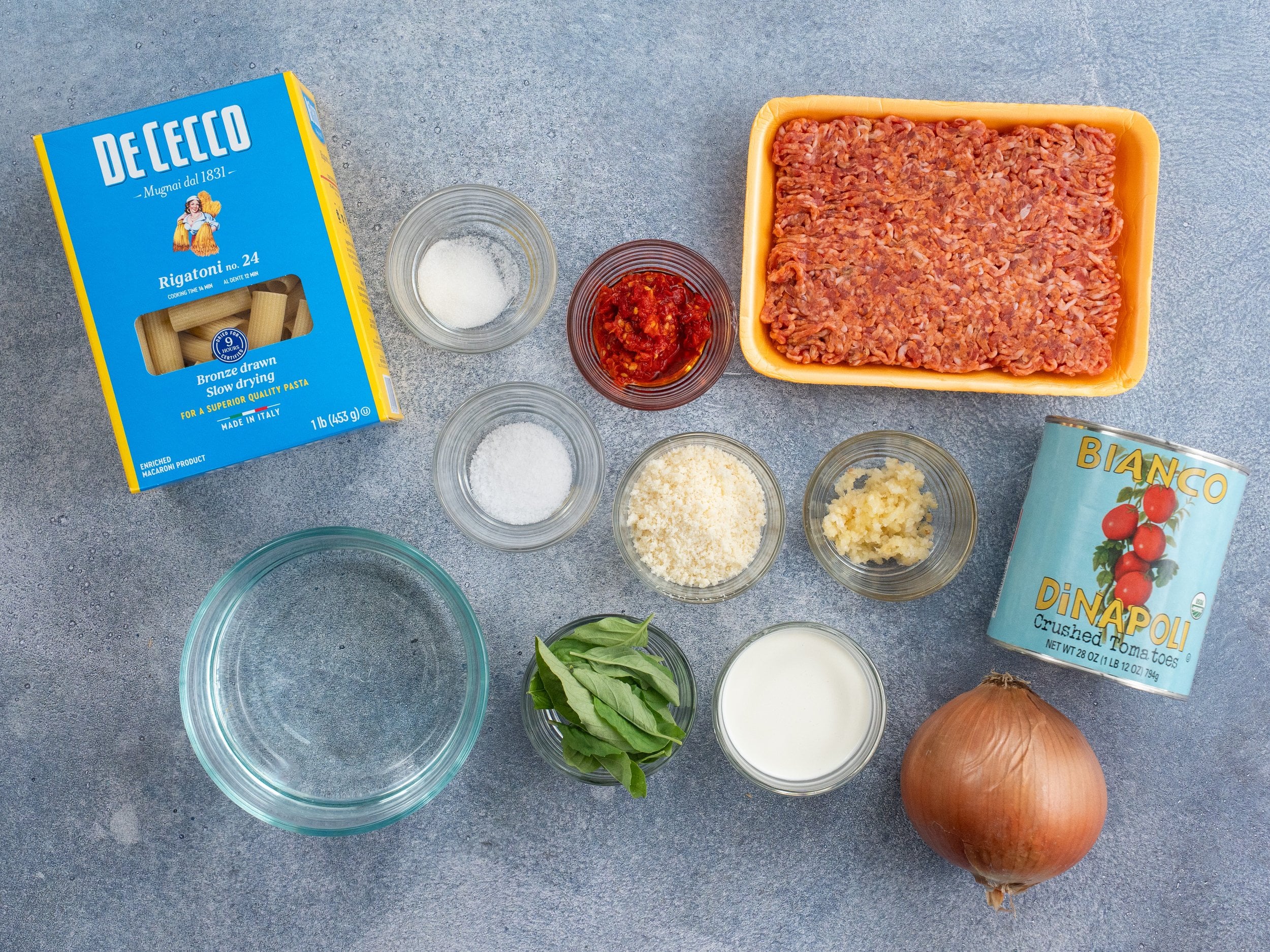 individual ingredients for making spicy sausage rigatoni pasta arranged on a grey background
