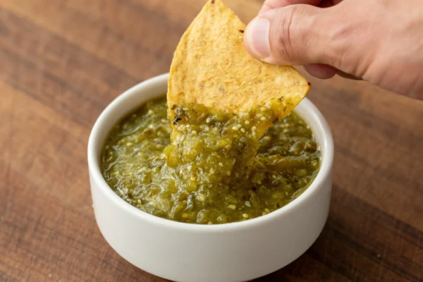 hand with tortilla chip dipping into white bowl of tomatillo salsa