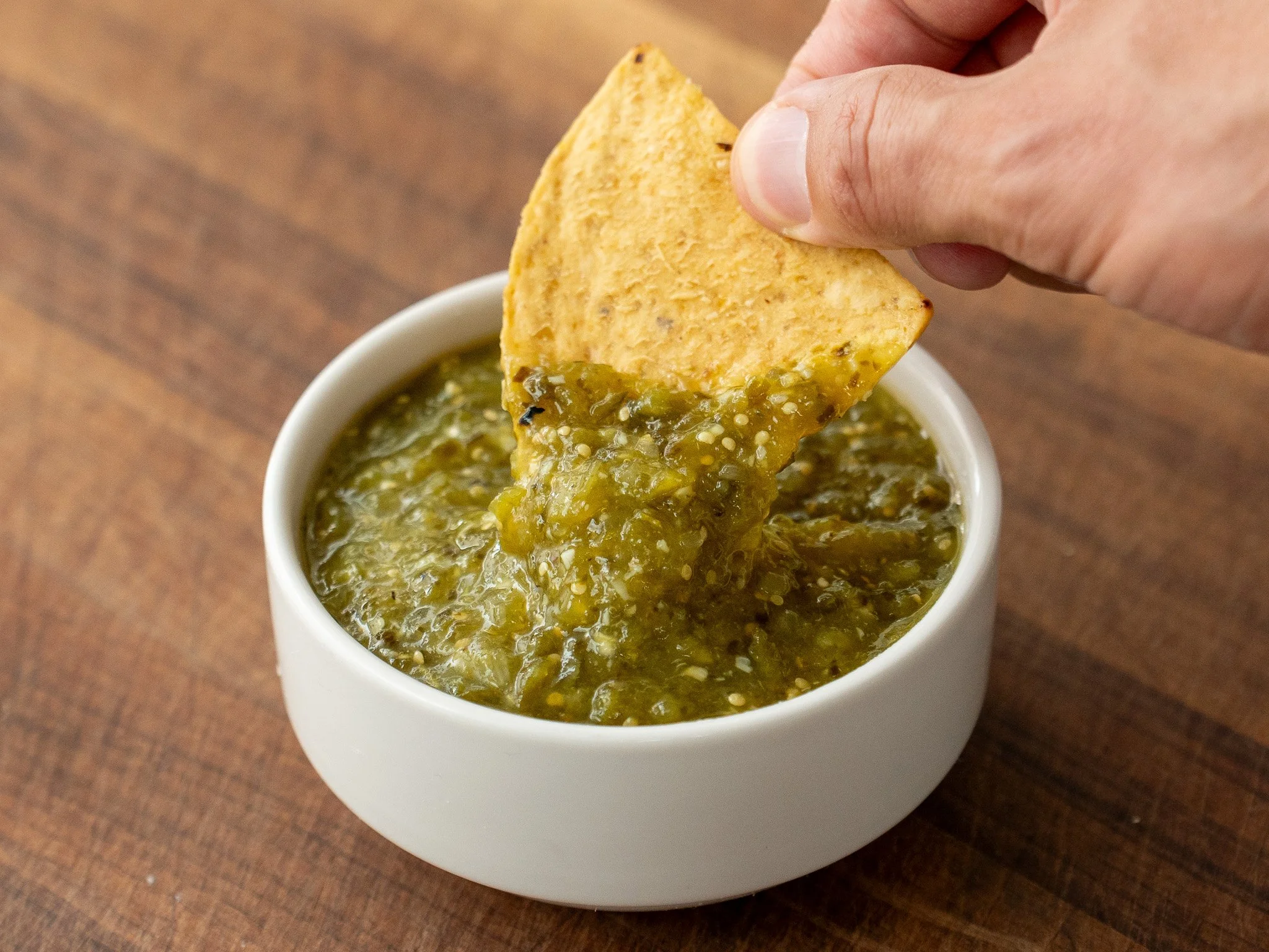 hand dipping tortilla chip into small white bowl tomatillo salsa