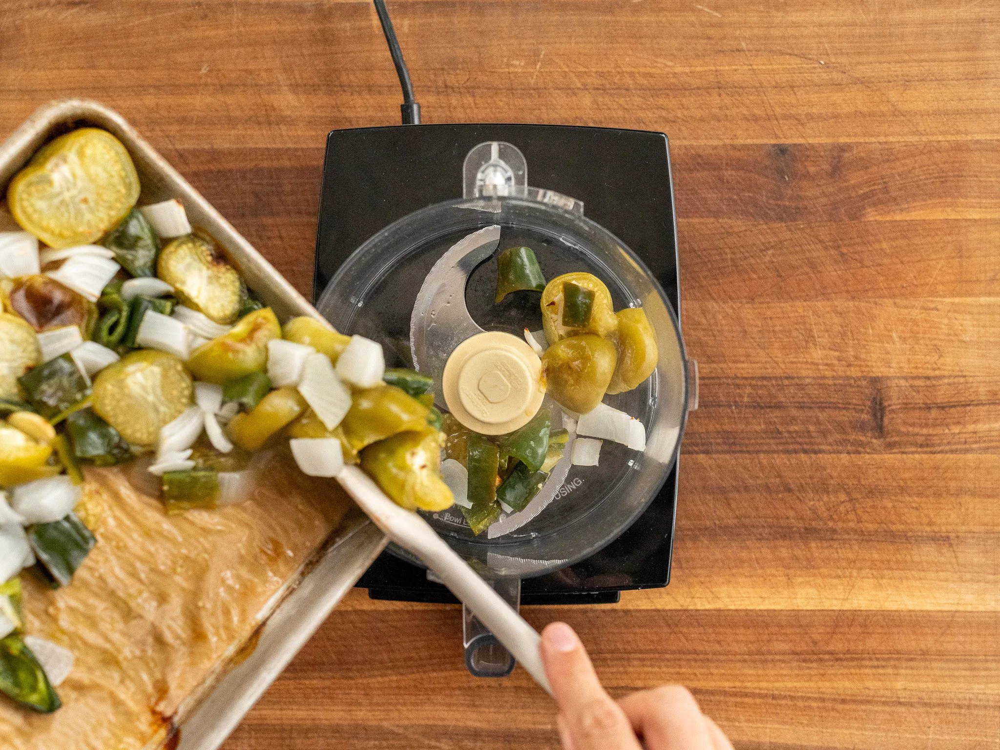 adding tray of roasted tomatillos onions peppers into food processor with white spatula