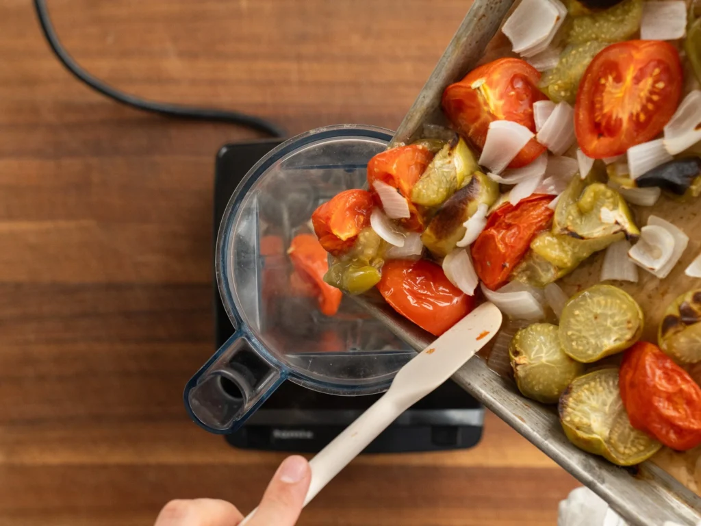 tomatoes and tomatillos being added to blender jar with white spatula