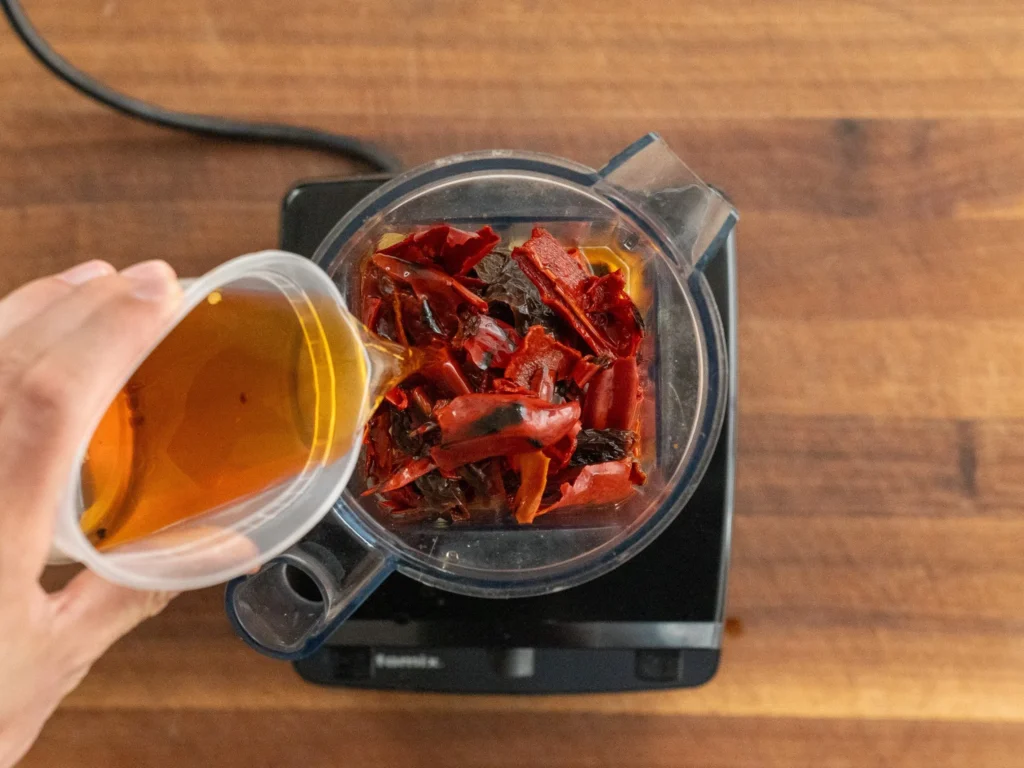 adding chili soaking water to blender jar with salsa ingredients