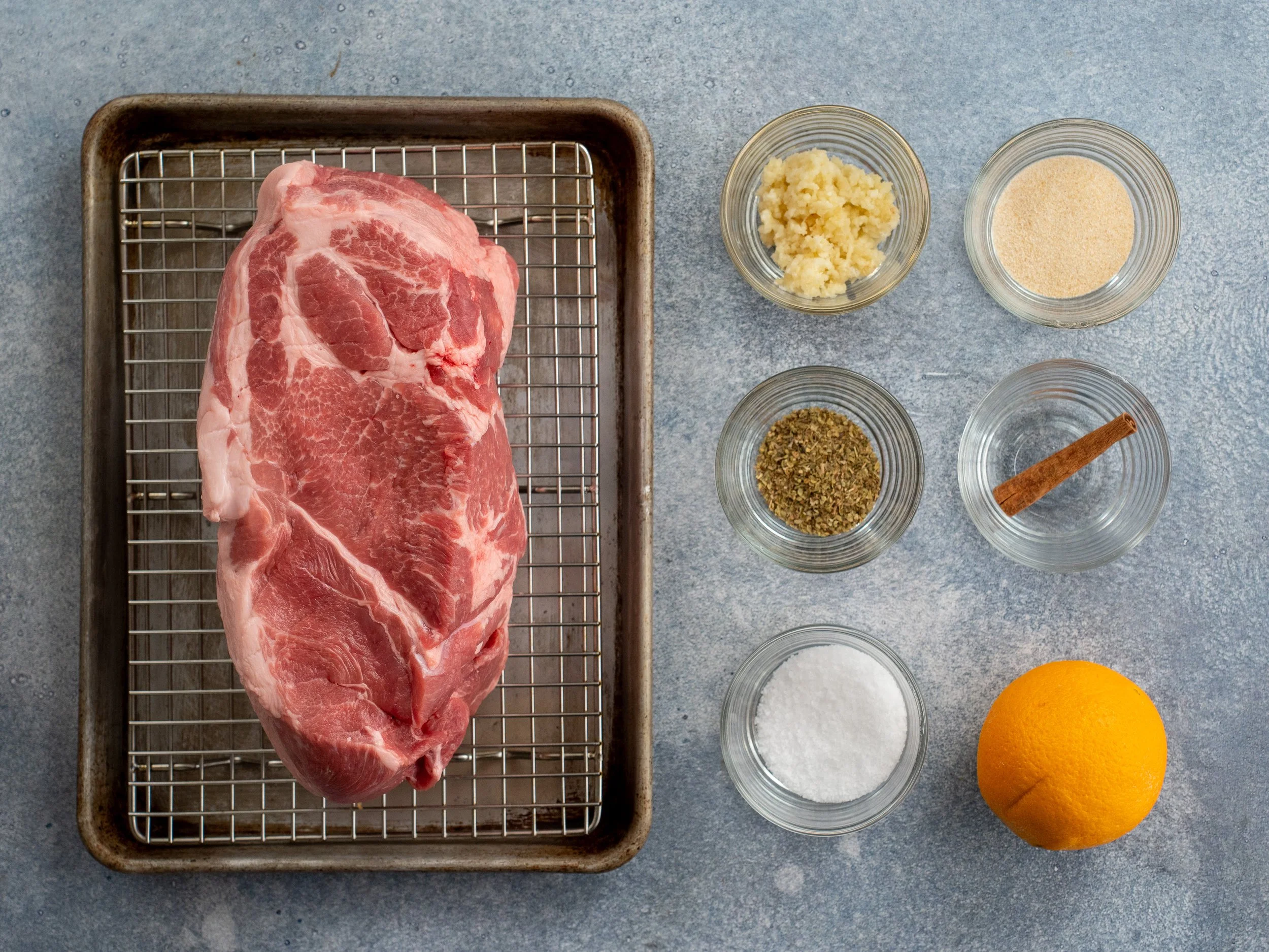 pork shoulder on sheet tray on the left with ingredients in glass bowls on the right side