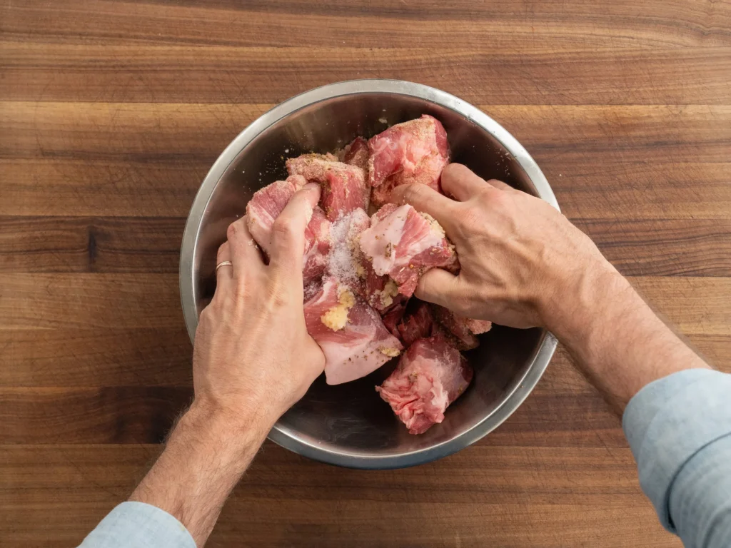 man mixing pork shoulder chunks with spices in a silver bowl