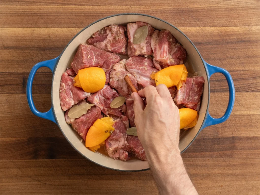 man's hand adding orange bay cinnamon into blue dutch oven filled with pork shoulder chunks