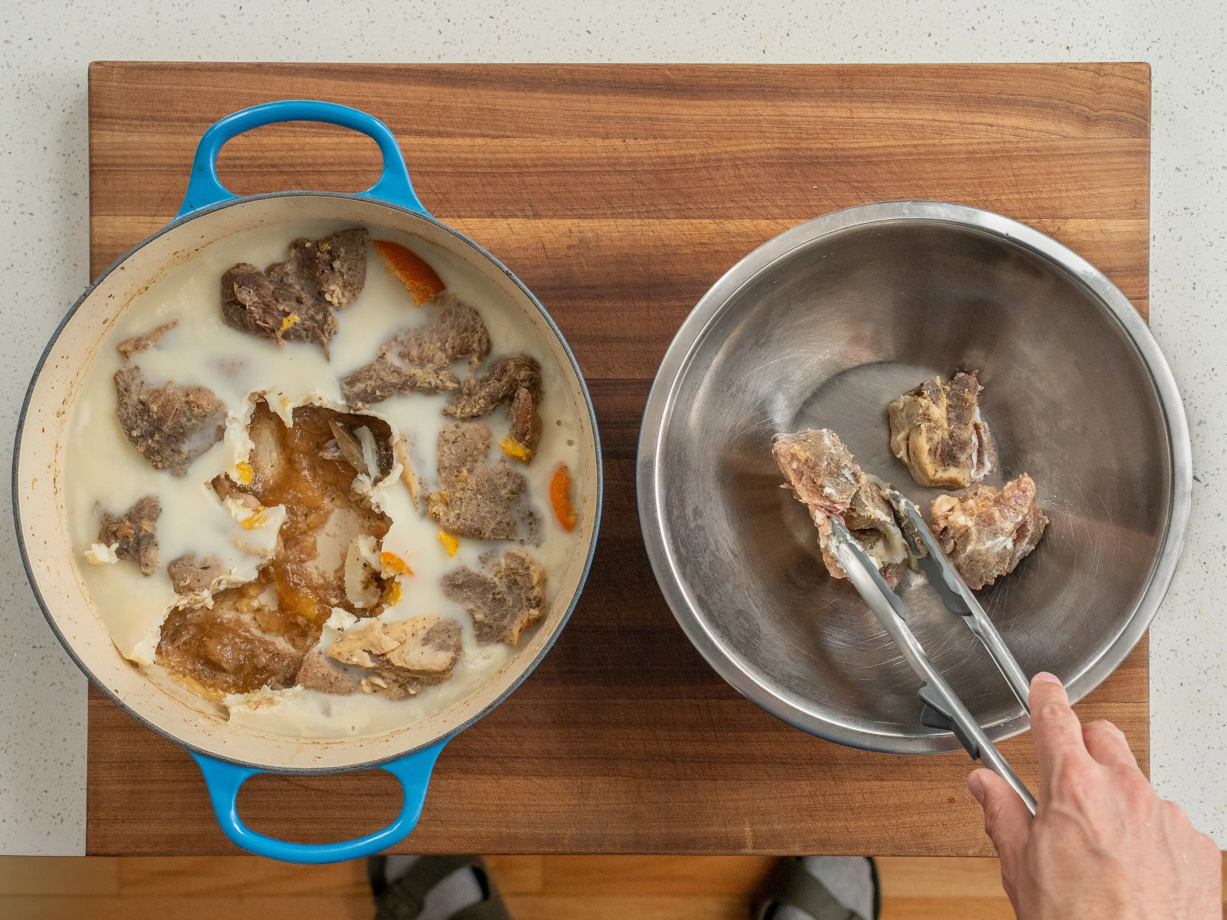 blue dutch oven filled with chilled pork shoulder in gelatinous juices on the right tongs removing chunks of pork shoulder from dutch oven and placing in metal bowl on right