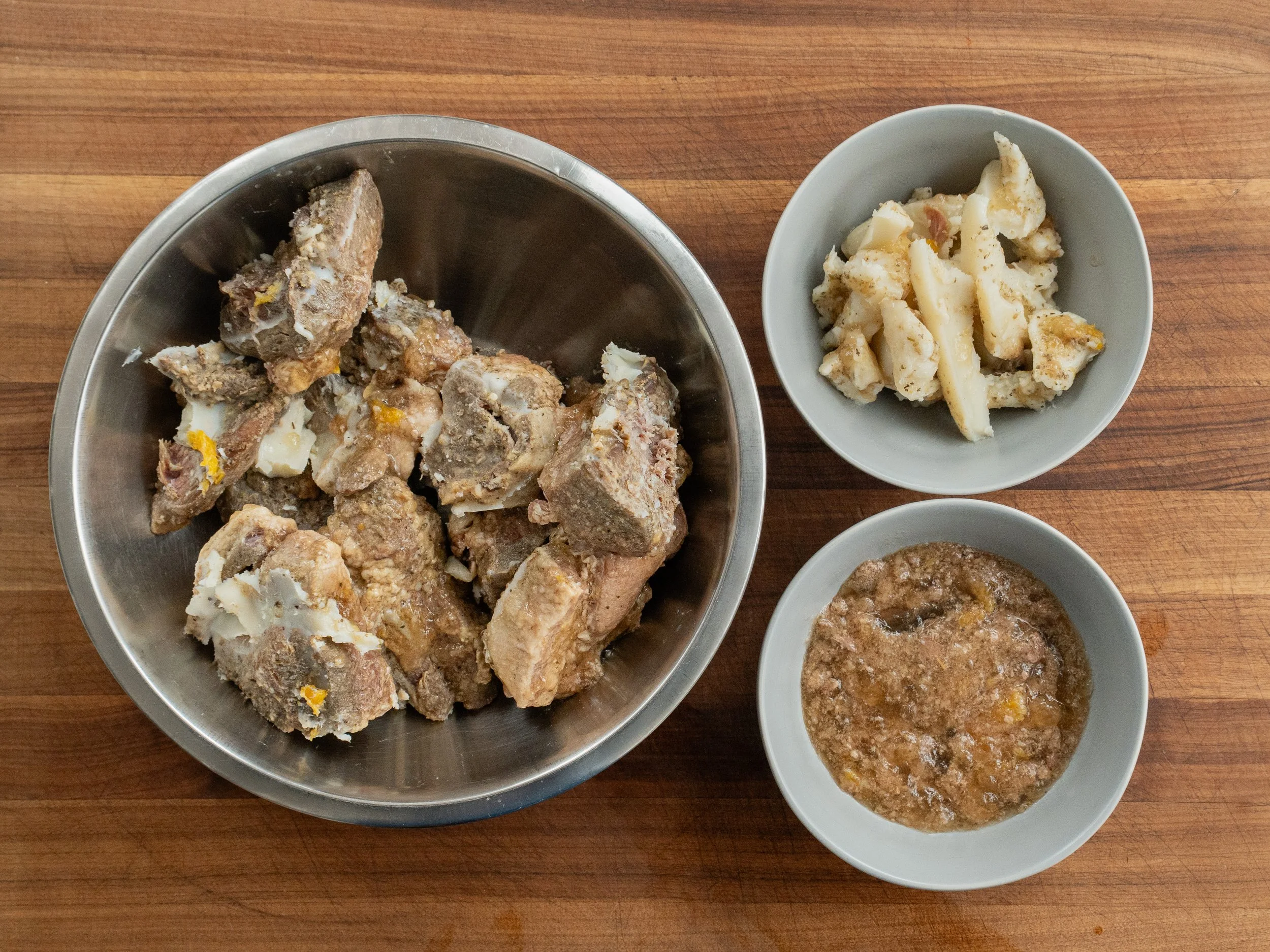 silver bowl of chilled cooked pork shoulder chunks on the left. grey bowl of pork fat on the upper right and grey bowl of pork juices on the lower right