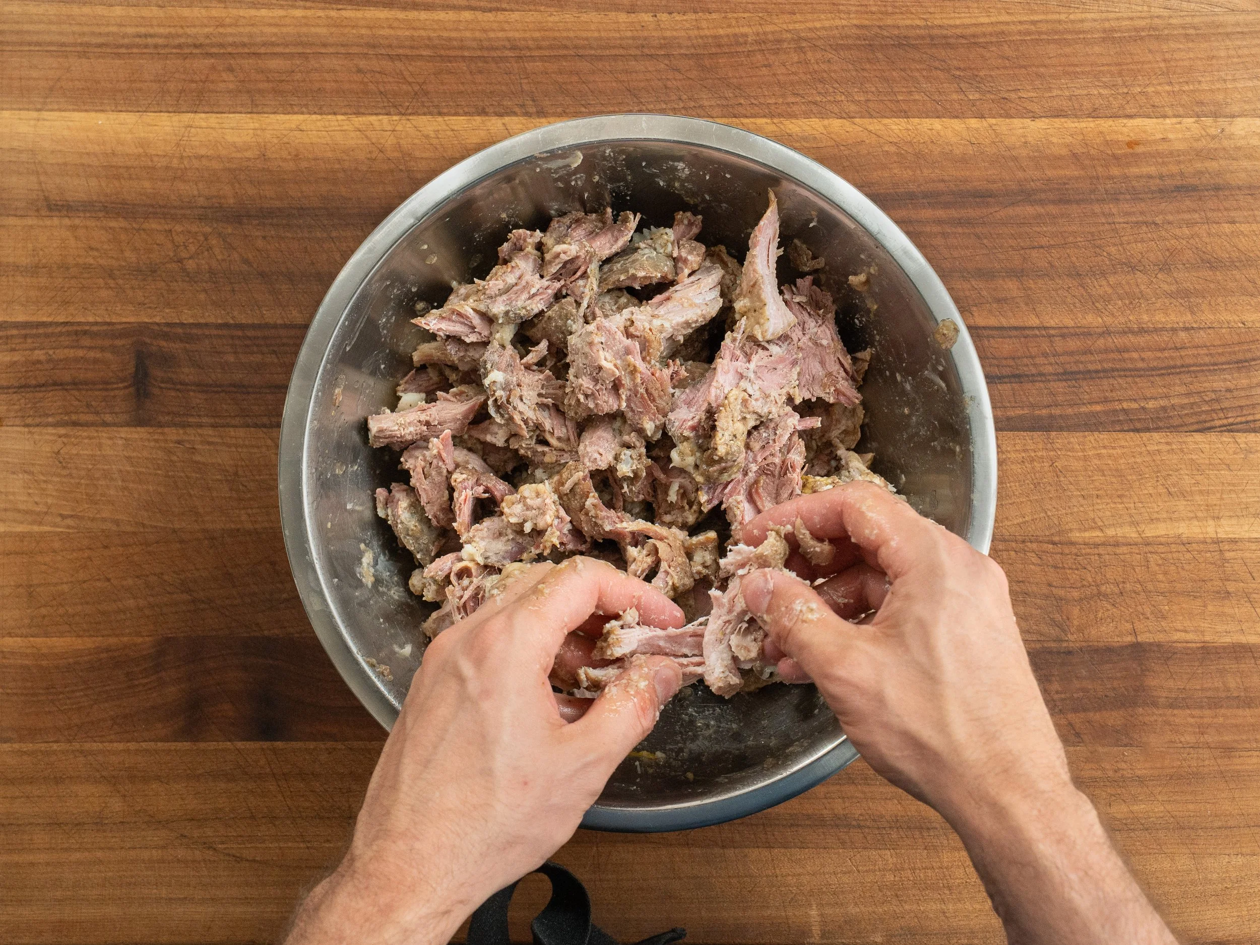 hands shredding apart pieces of pork in a metal bowl