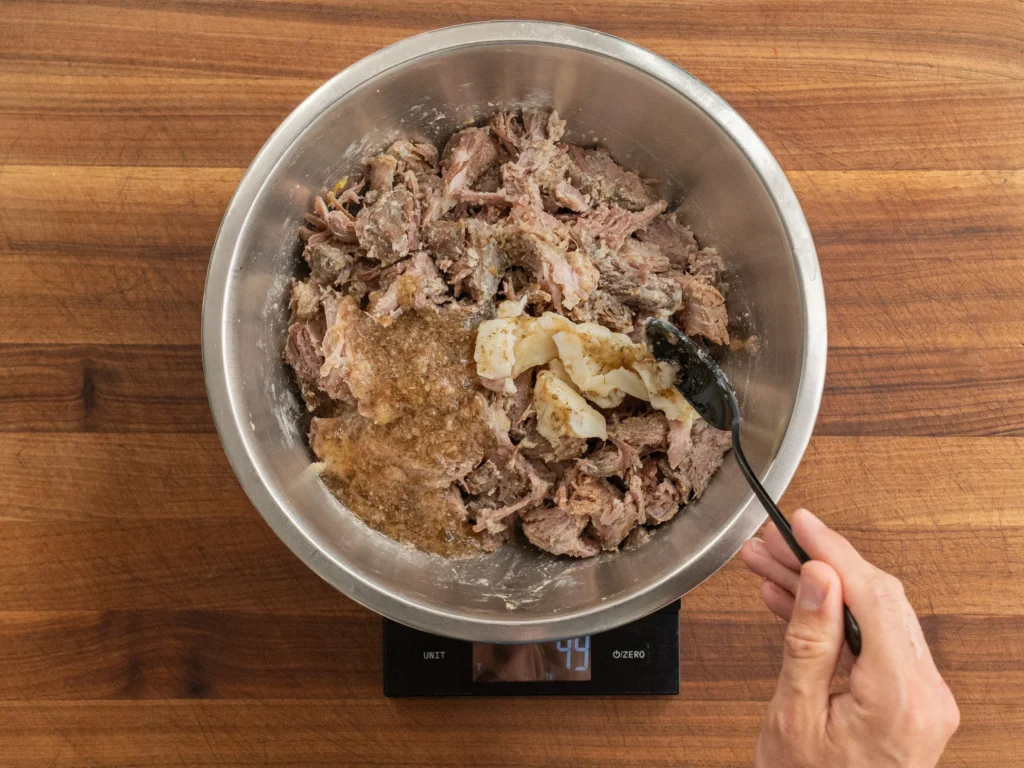 black spoon adding pork stock and fat into silver bowl filled with carnitas