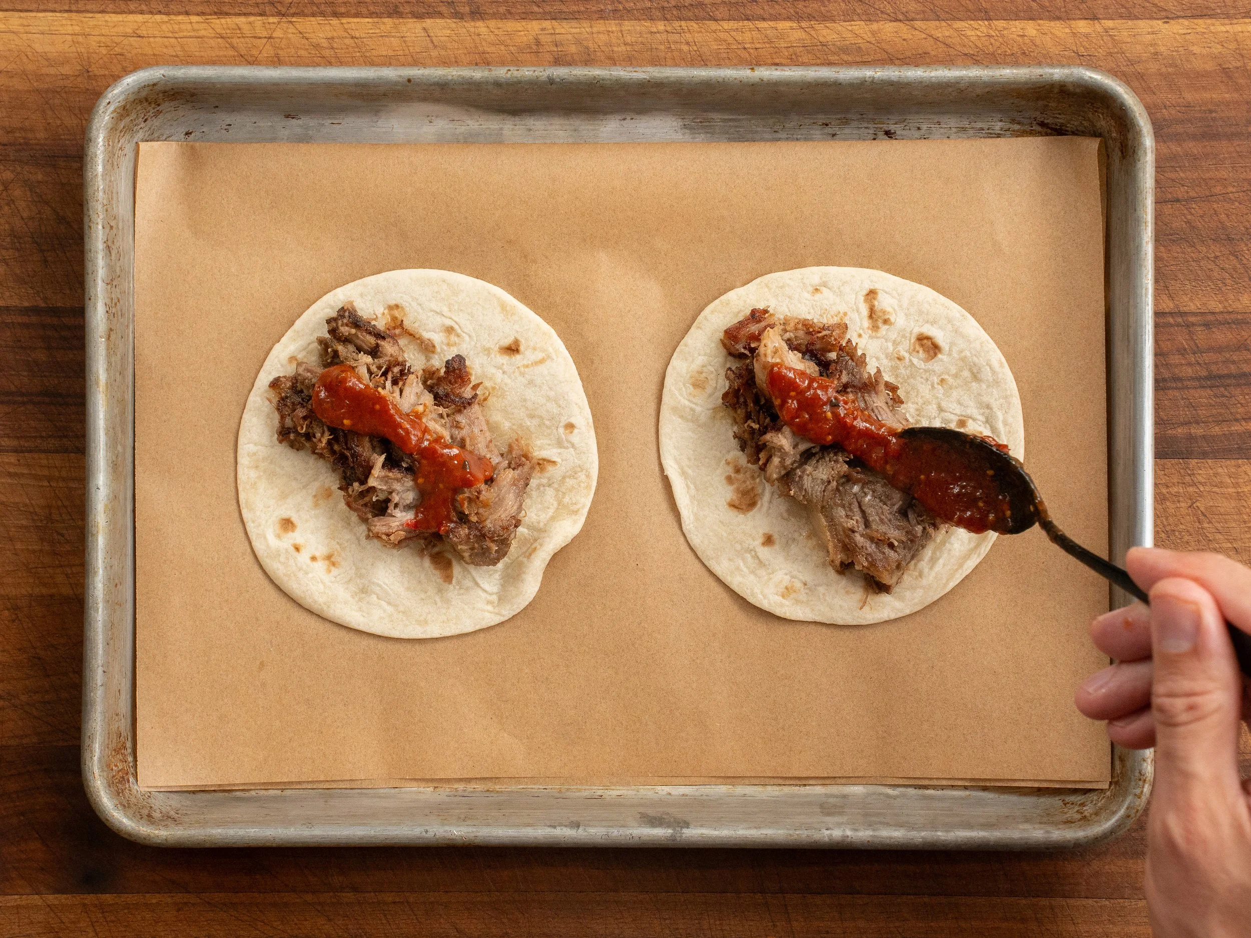 black spoon placing red salsa on flour tortillas with carnitas on a sheet tray