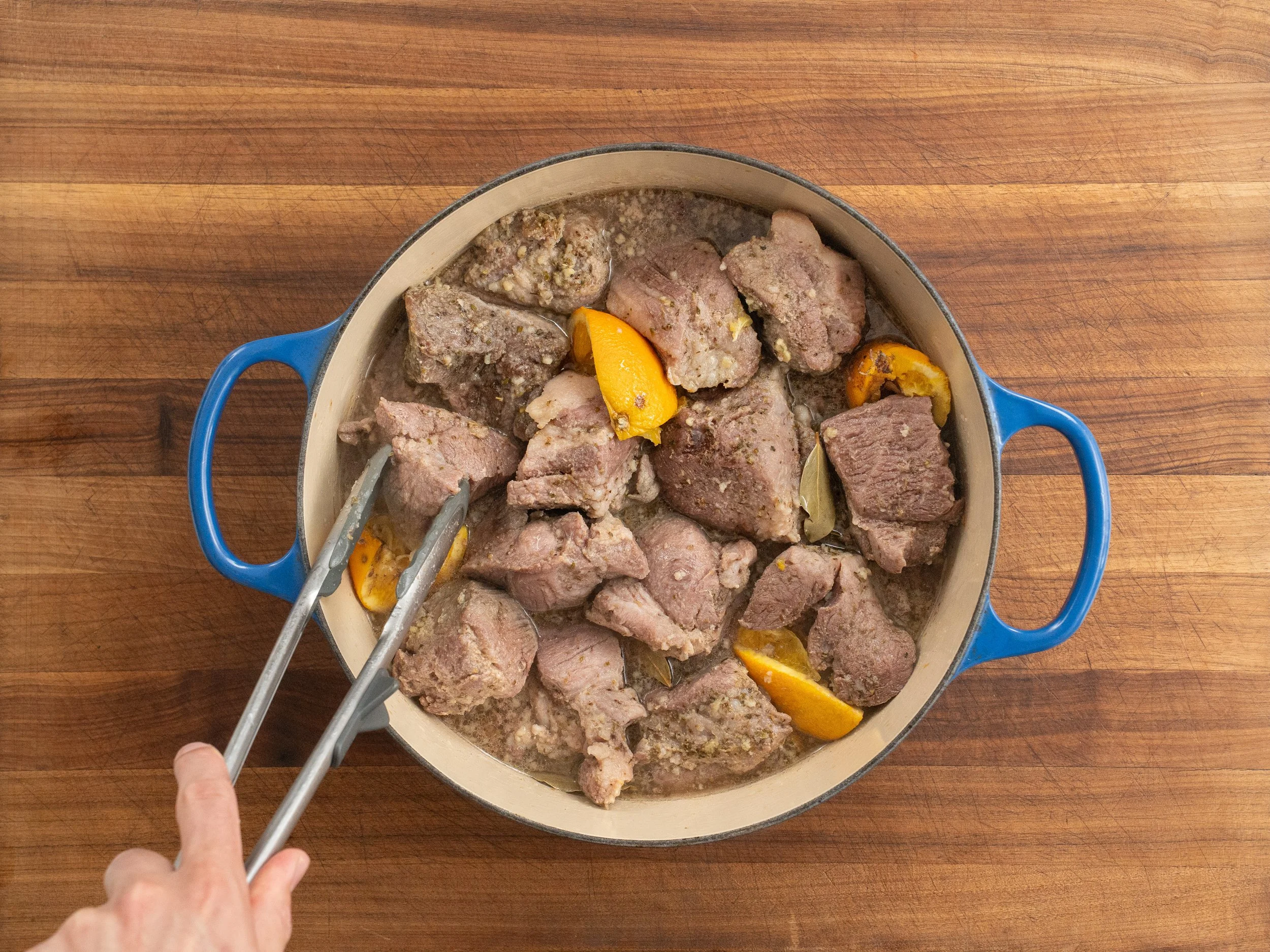 tongs flipping pieces of cooking pork shoulder in blue dutch oven