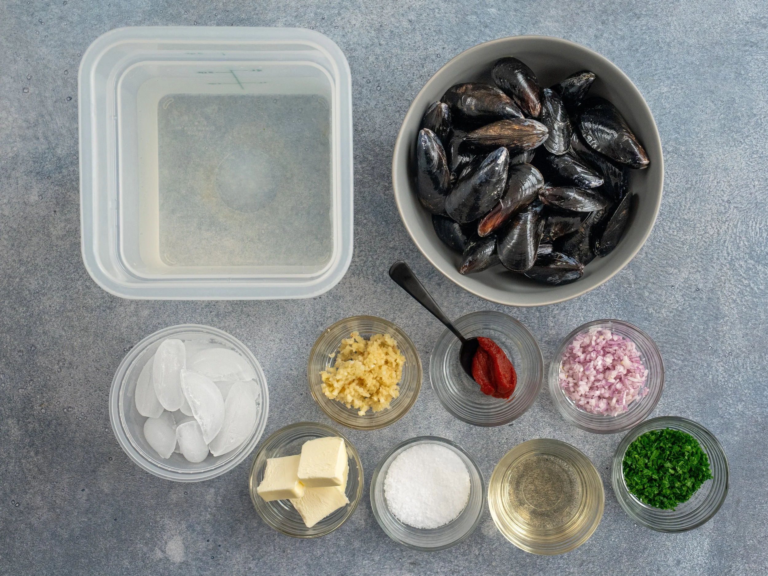 container of water, bowl of mussels, ice, garlic, butter, tomato paste, shallots, wine, parsley in bowls on grey background