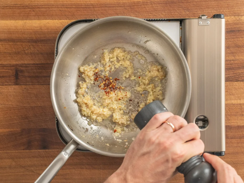 using pepper grinder to add pepper to silver pot of simmering garlic shallot chili flake