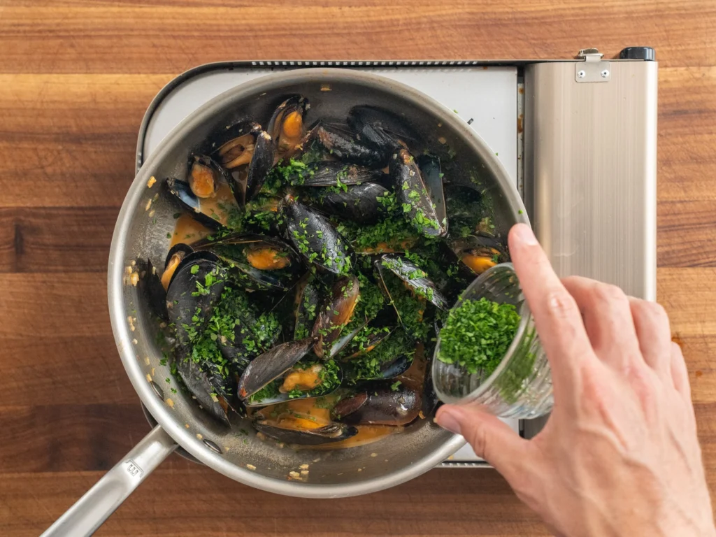 adding cup of chopped parsley into silver pan filled with cooked mussels in white wine sauce