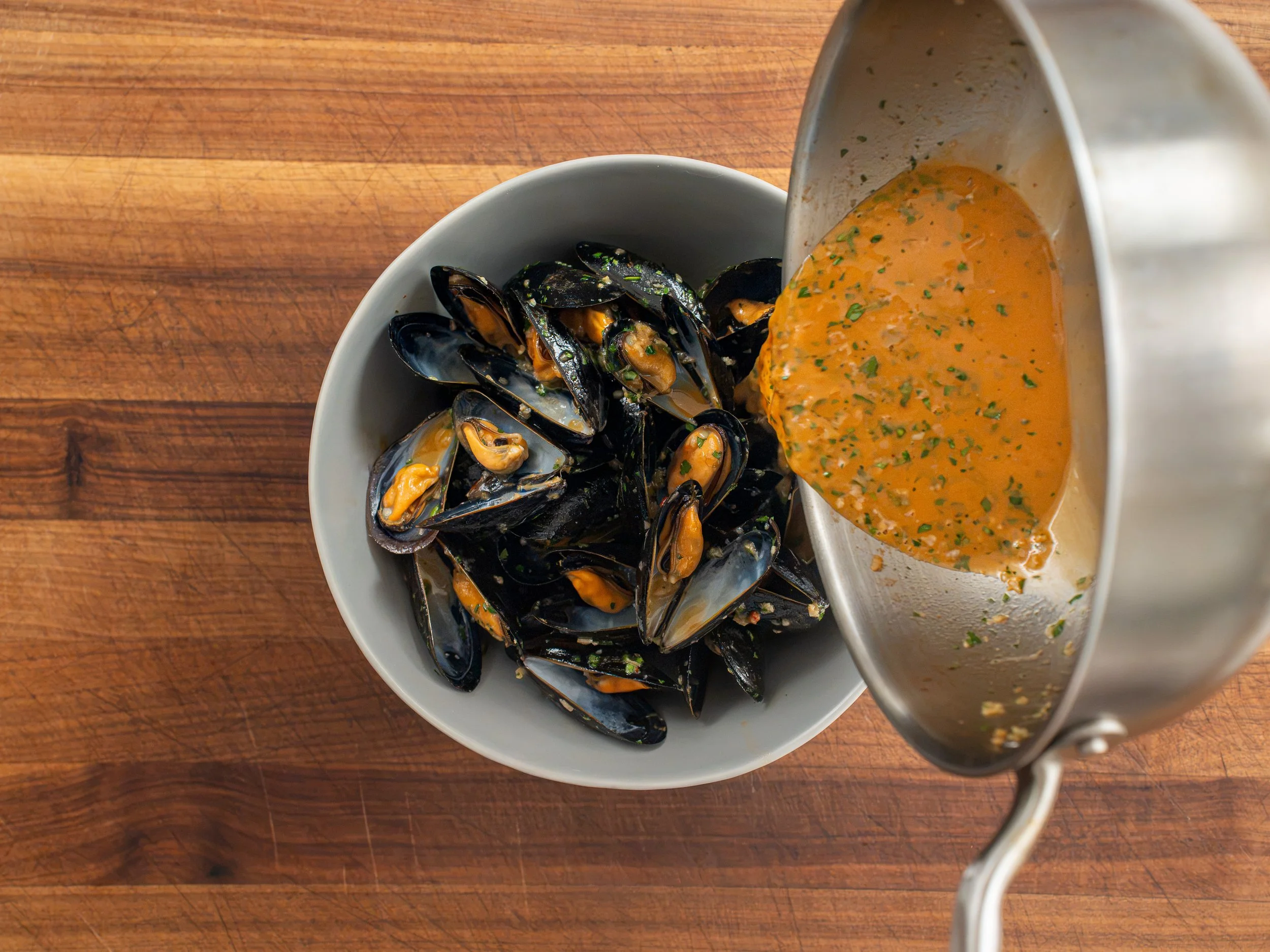 white wine sauce in silver pot being poured into grey bowl filled with cooked mussels