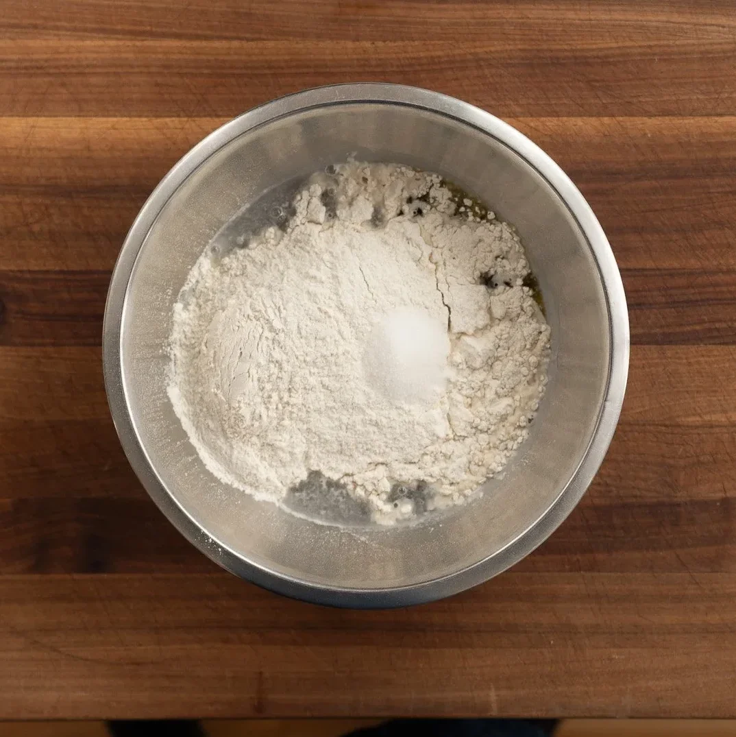 flour water salt and yeast in a silver mixing bowl on a brown cutting board
