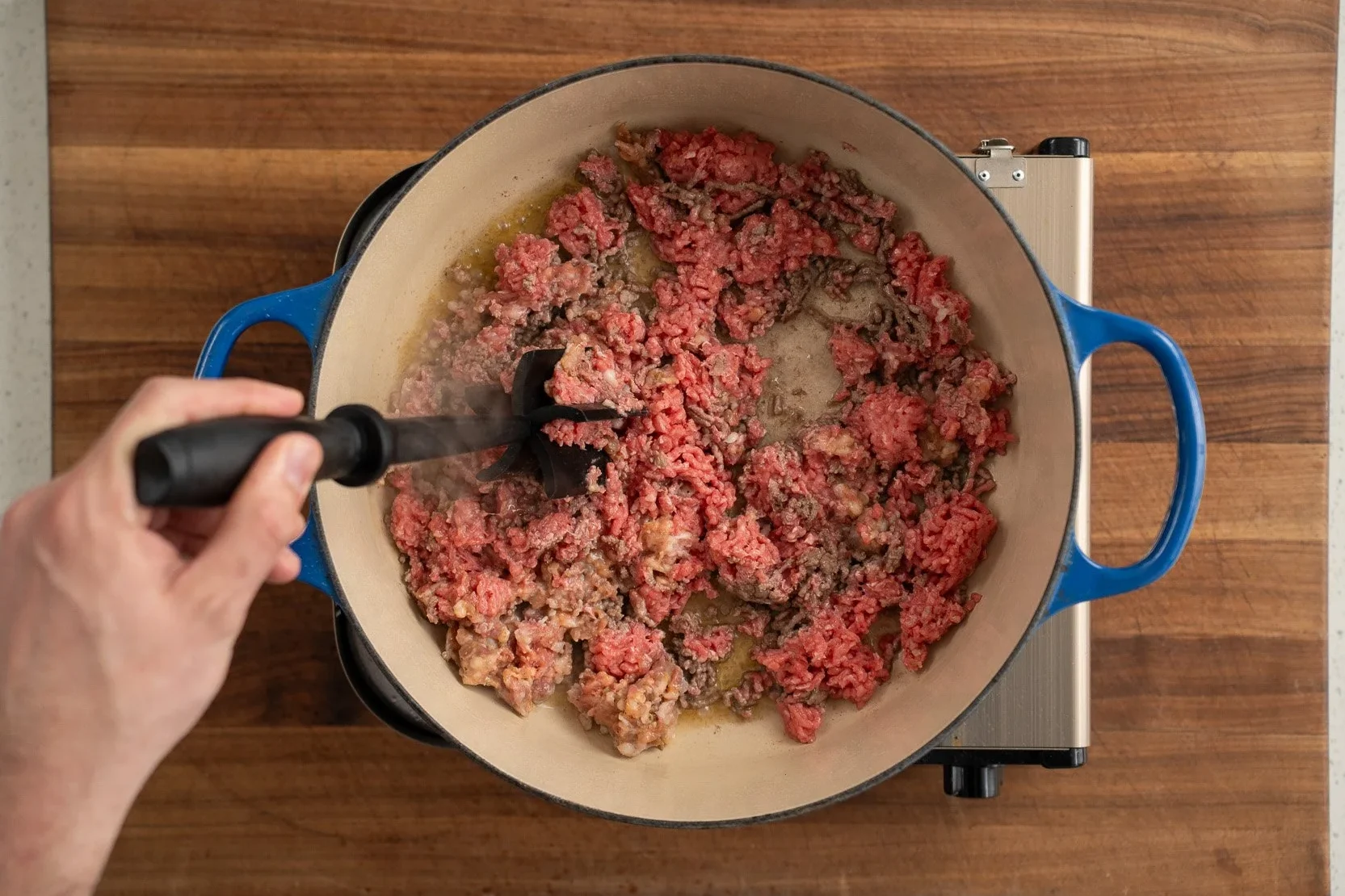 breaking down ground beef with a black meat chopper in a blue dutch oven