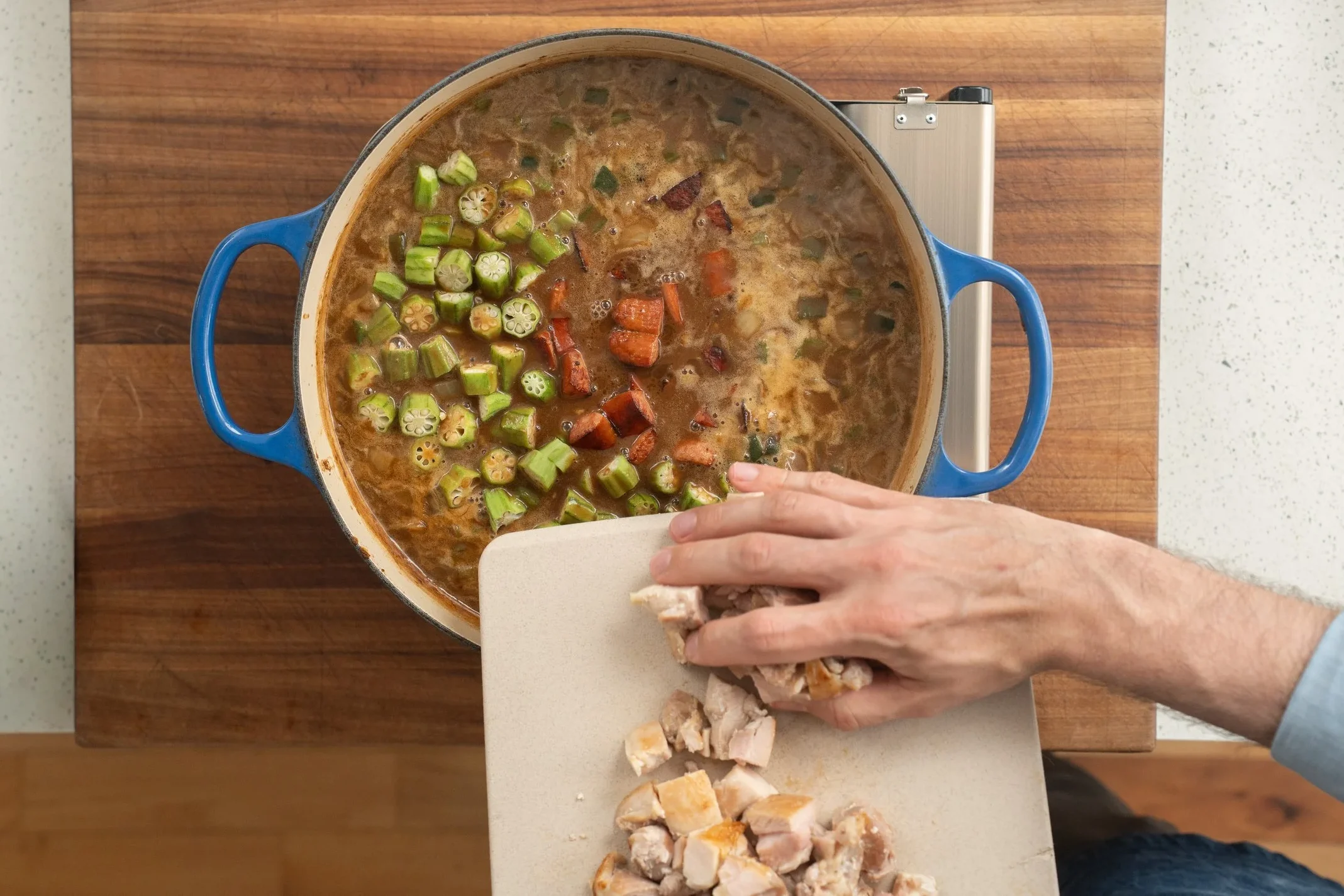 adding chicken thighs andouille and okra to a blue dutch oven filled with gumbo