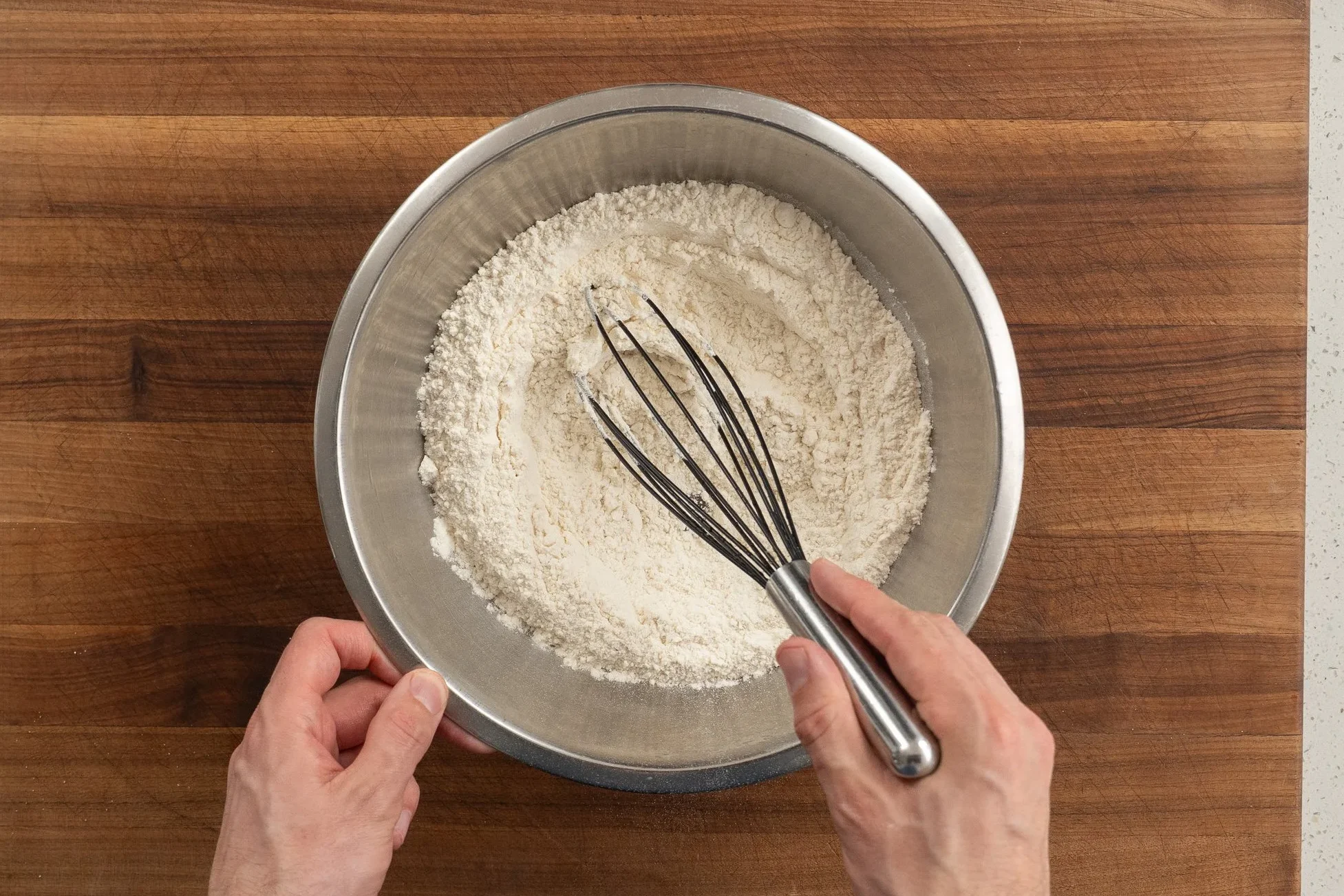 whisking flour baking soda and salt in a metal bowl on a brown cutting board