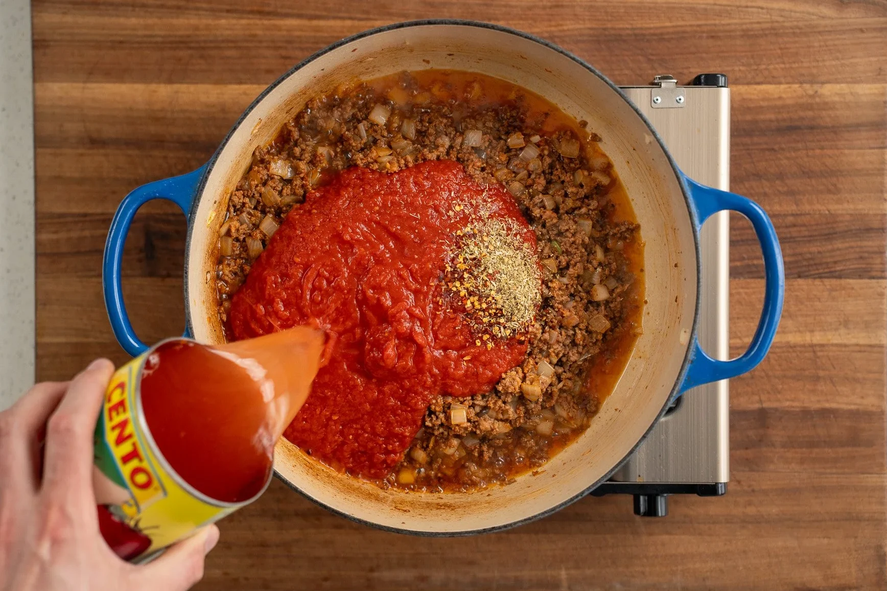 adding crushed tomatoes and spices to a blue dutch oven