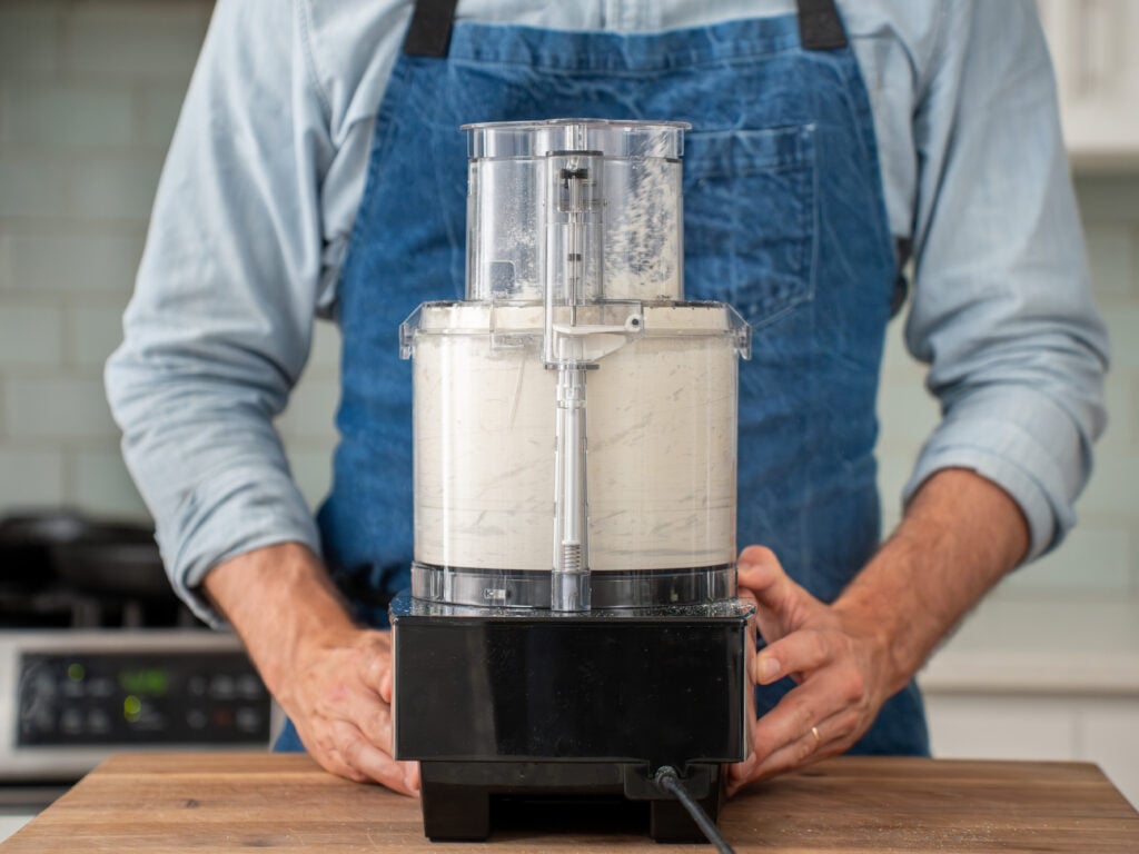 man in blue apron standing behind food processor filled with dry biscuit ingredients