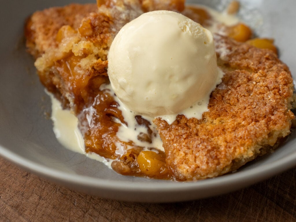 grey bowl filled with portion of peach cobbler topped with scoop of vanilla ice cream