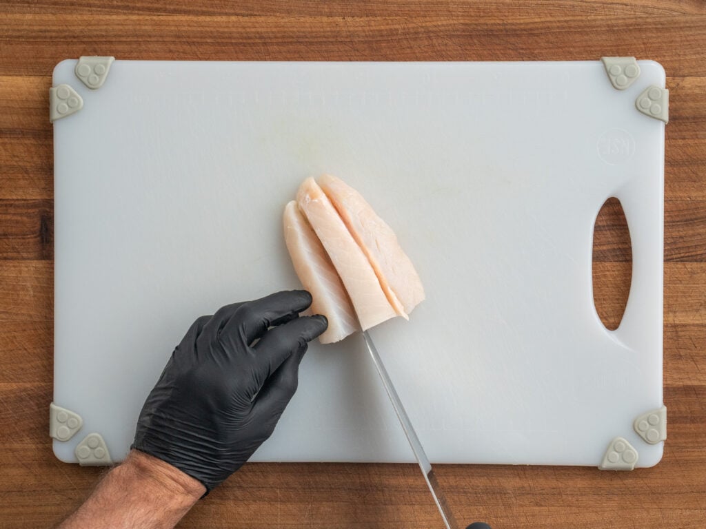 black gloved hand slicing Atlantic cod into planks on top of white plastic cutting board