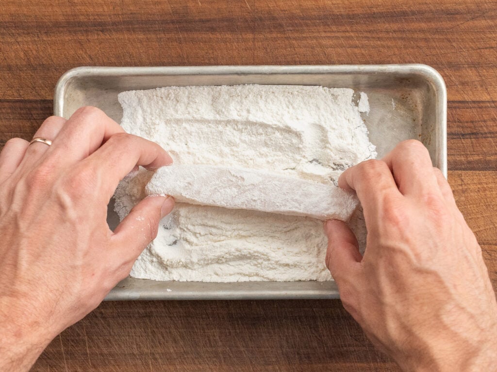 using fingers to dredge plank of Atlantic cod in flour on small silver sheet tray