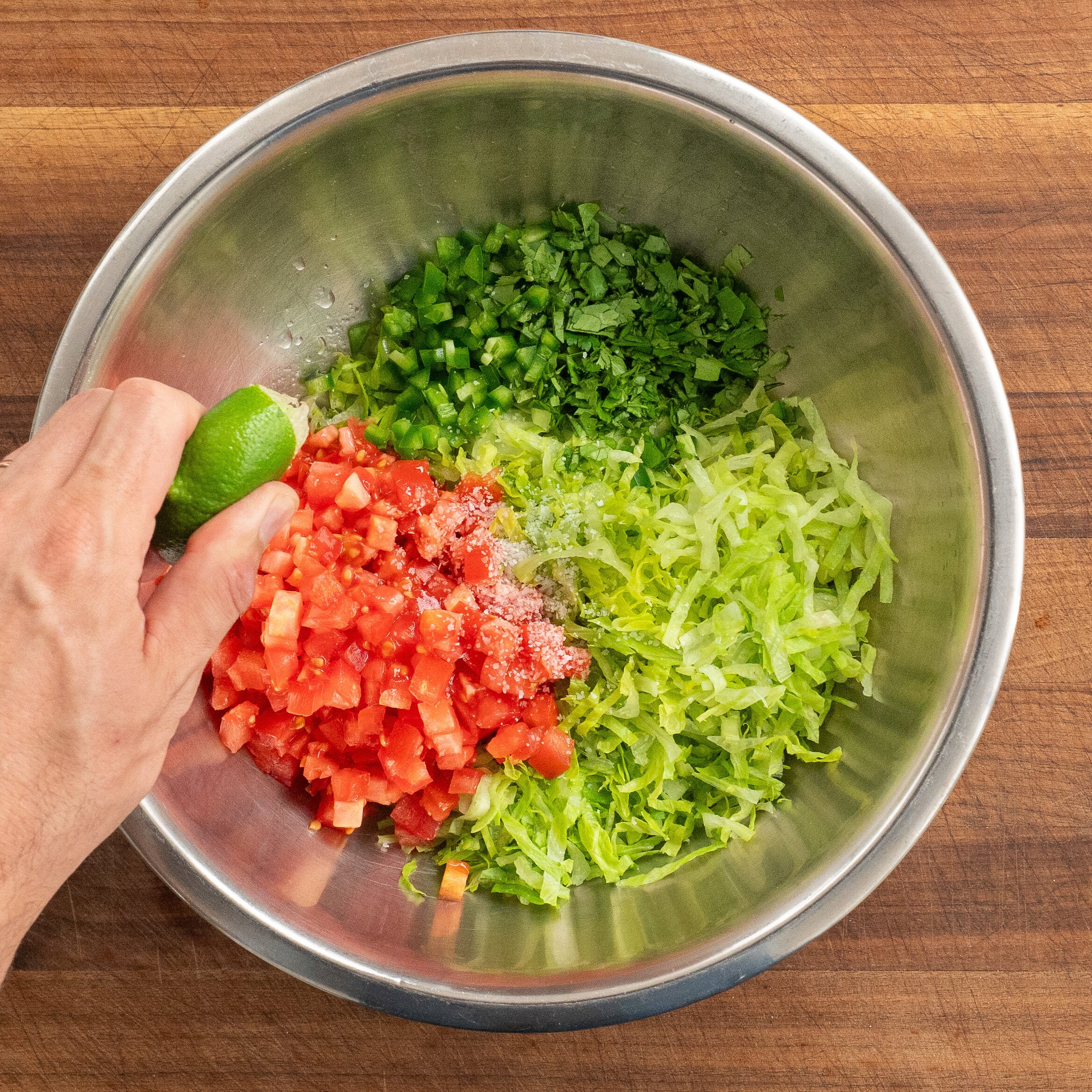 hand squeezing lime juice into silver bowl filled with shredded lettuce cilantro tomato