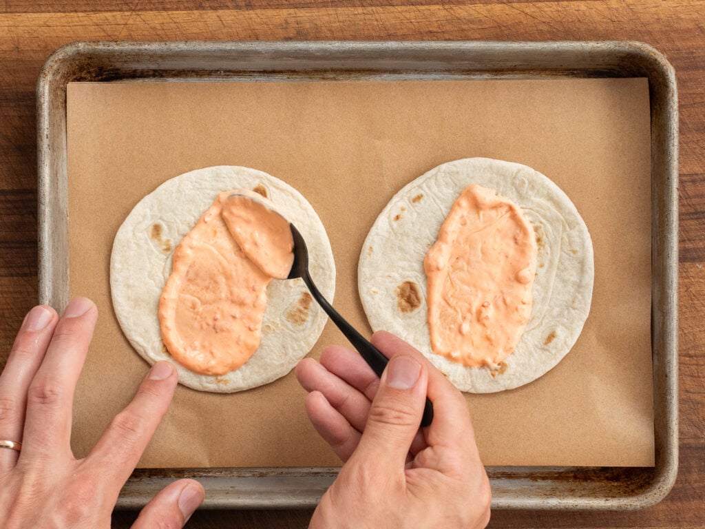 hands using black spoon to spread sambar mayo onto 2 small corn tortillas