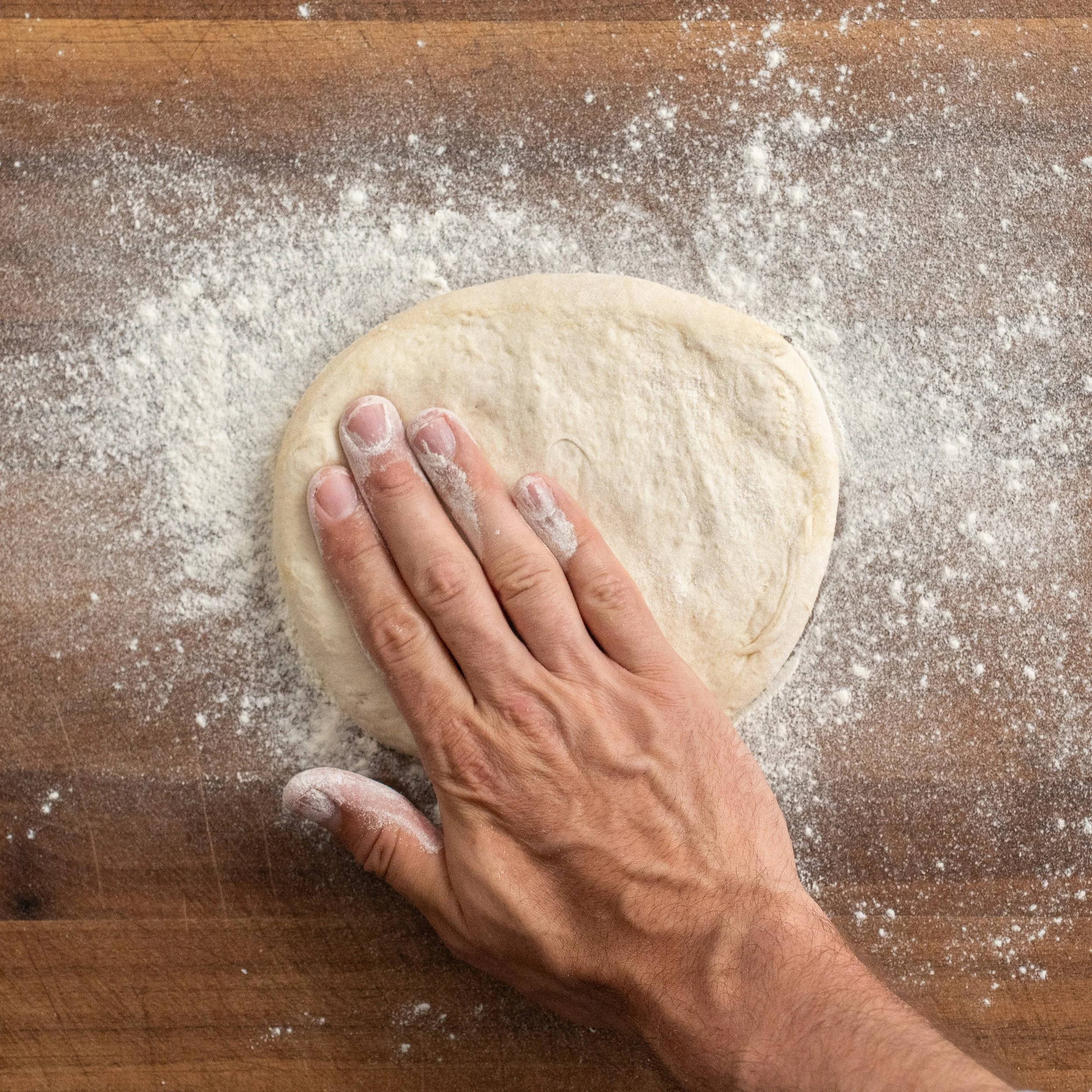 hand patting down floured ball of pizza dough on a wooden surface