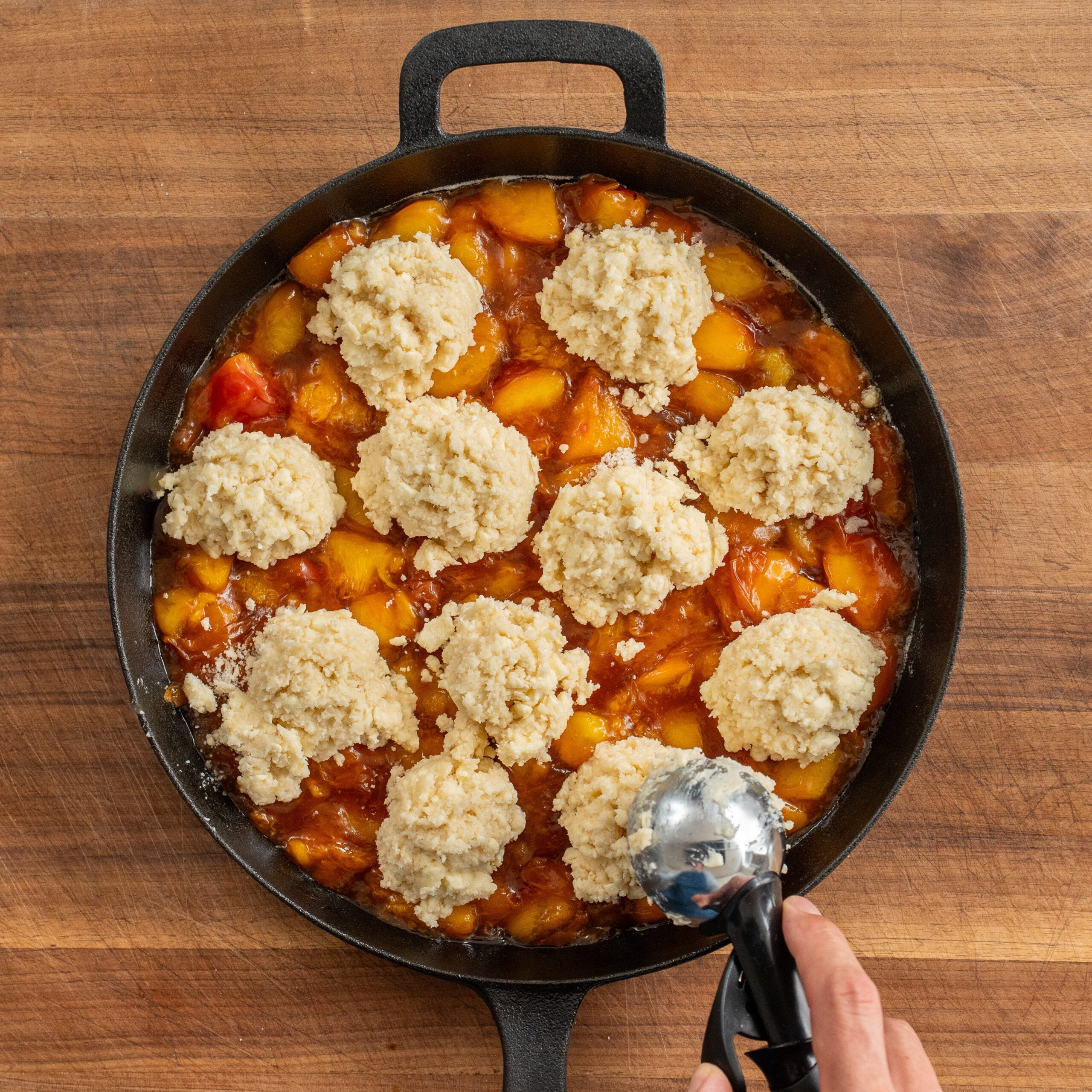 using a scoop to add drop biscuits to cast iron pan filled with cooked peach filling